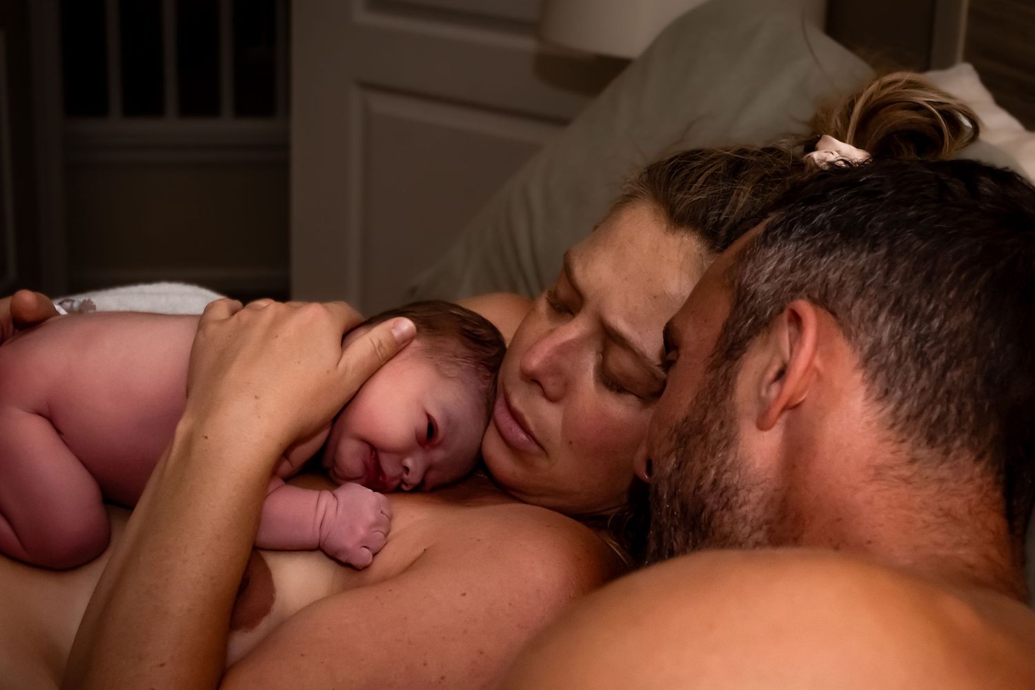 Image of a mother and father snuggling while the mother holds their newborn baby on her chest.