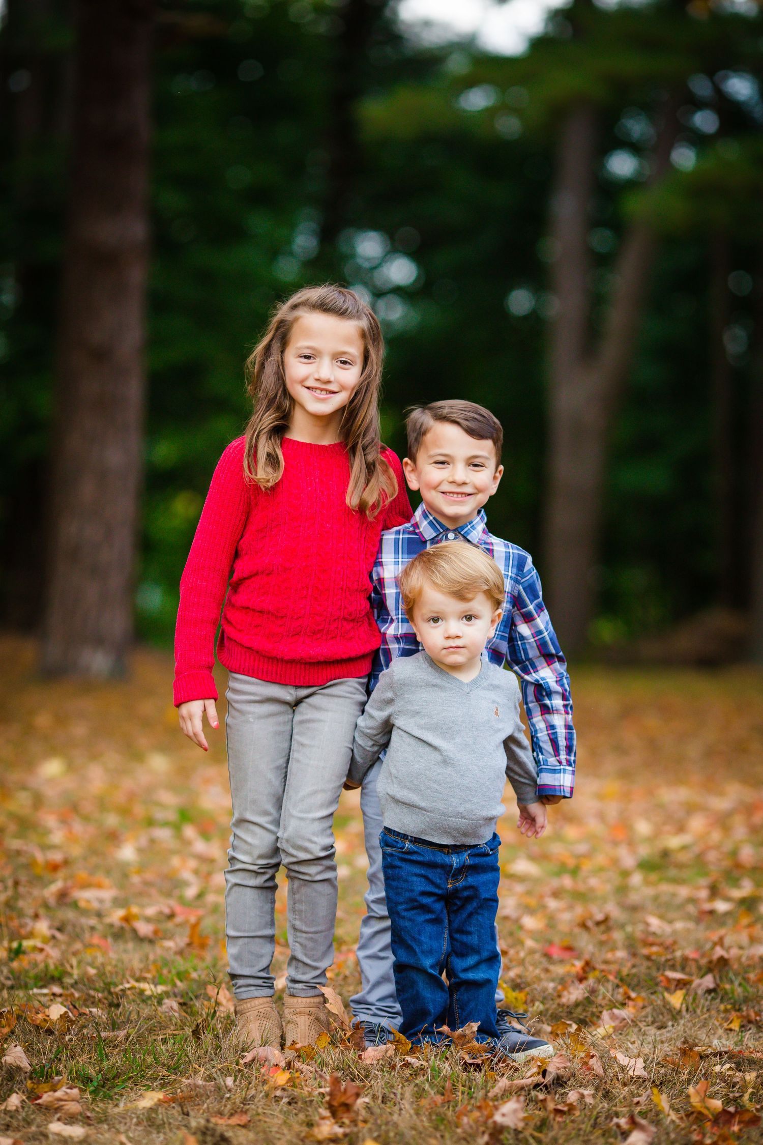 Three siblings standing together in autumn forest wearing casual clothing.