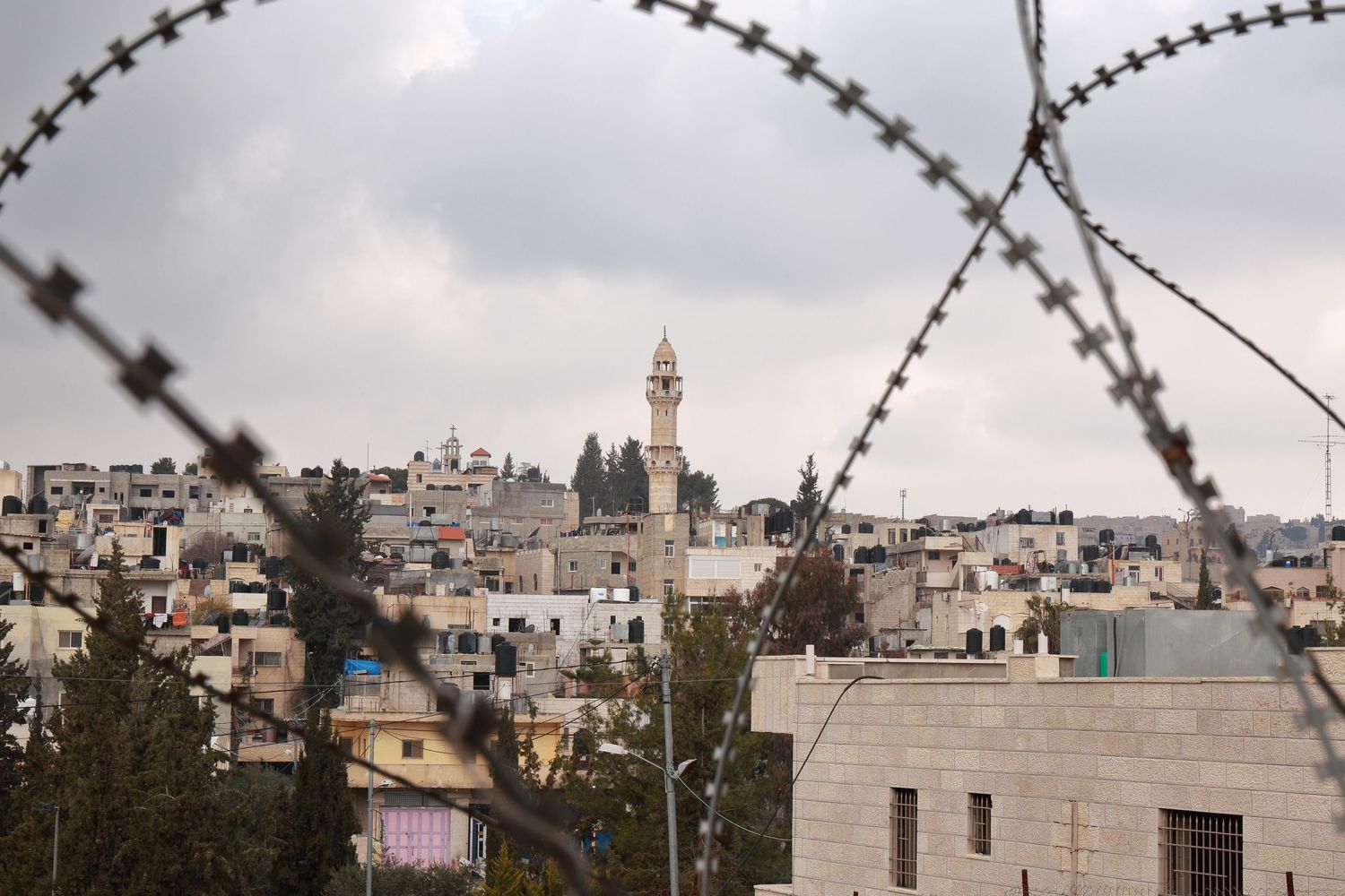Cityscape view through barbed wire showing a minaret and buildings in a Middle Eastern town.