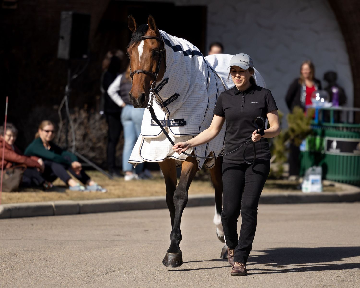 Handler walking beside a brown horse wearing a white blanket at an equestrian event.