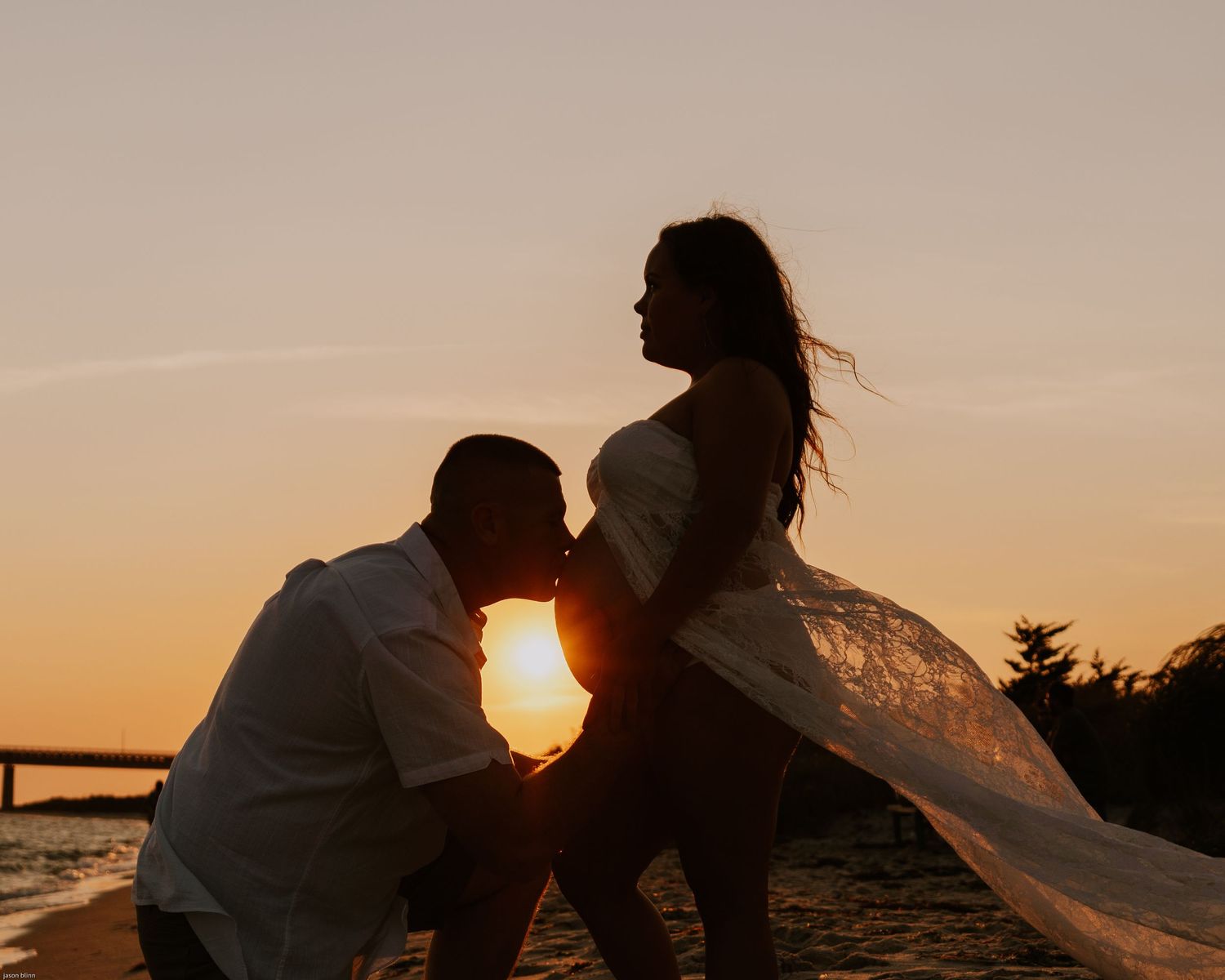 Maternity beach photo with flowing dress silhouetted against sunset.