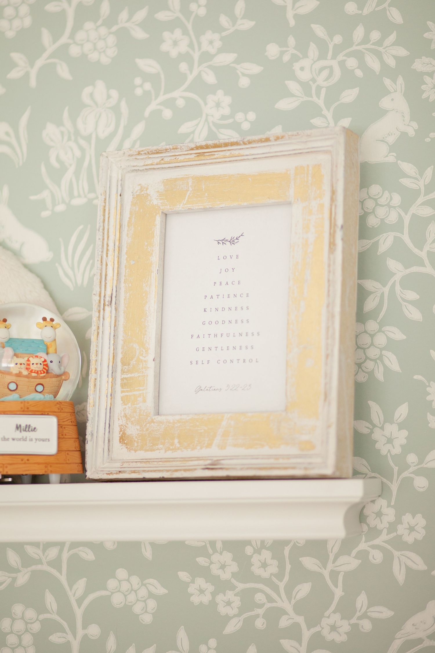 A distressed white picture frame with yellow accents sits on a shelf against mint floral wallpaper.