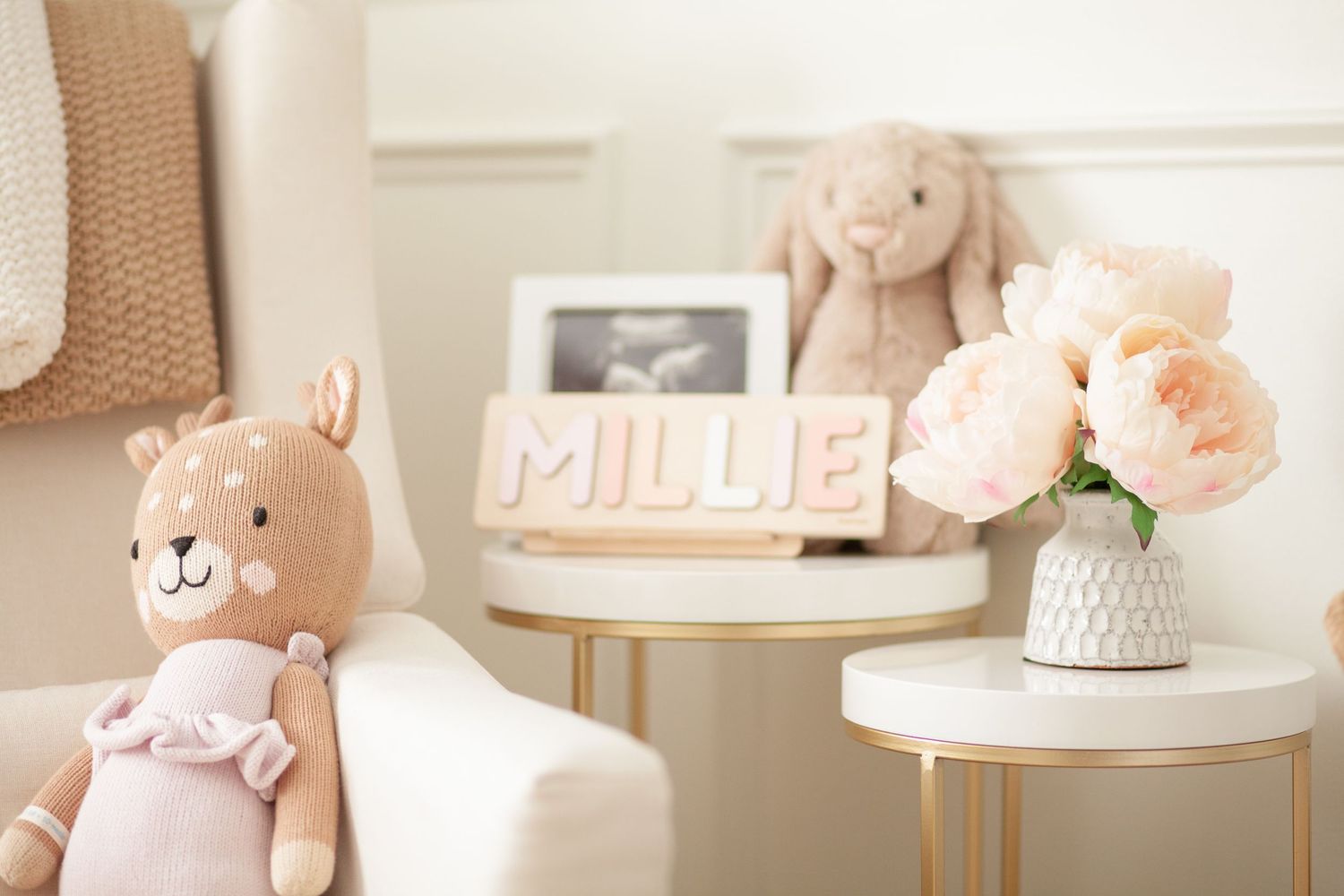 Soft pink nursery decor with plush bunny toy, framed sign, and peach roses in white vase on gold side table.