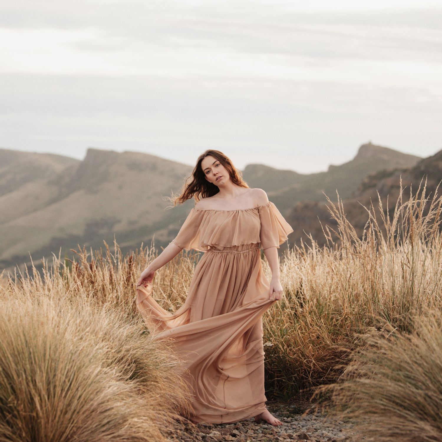 A person in a flowing peach-colored dress poses gracefully among tall grasses on a hillside.