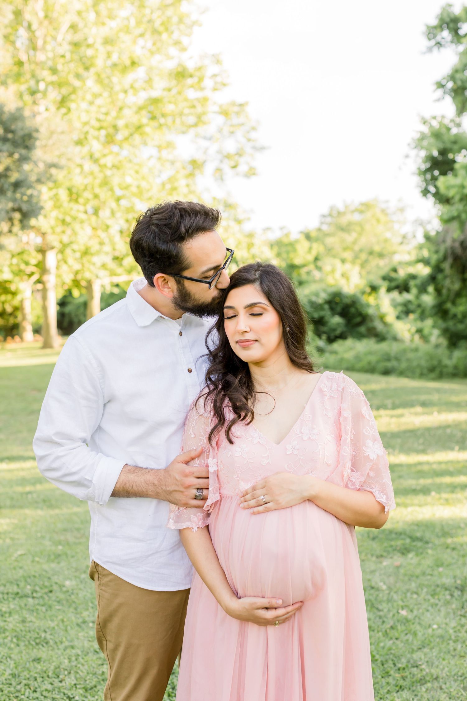 Intimate maternity portrait of couple embracing in garden with woman in pink flowing dress.