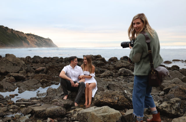 Coastal photoshoot scene with people and camera equipment on rocky beach at sunset.