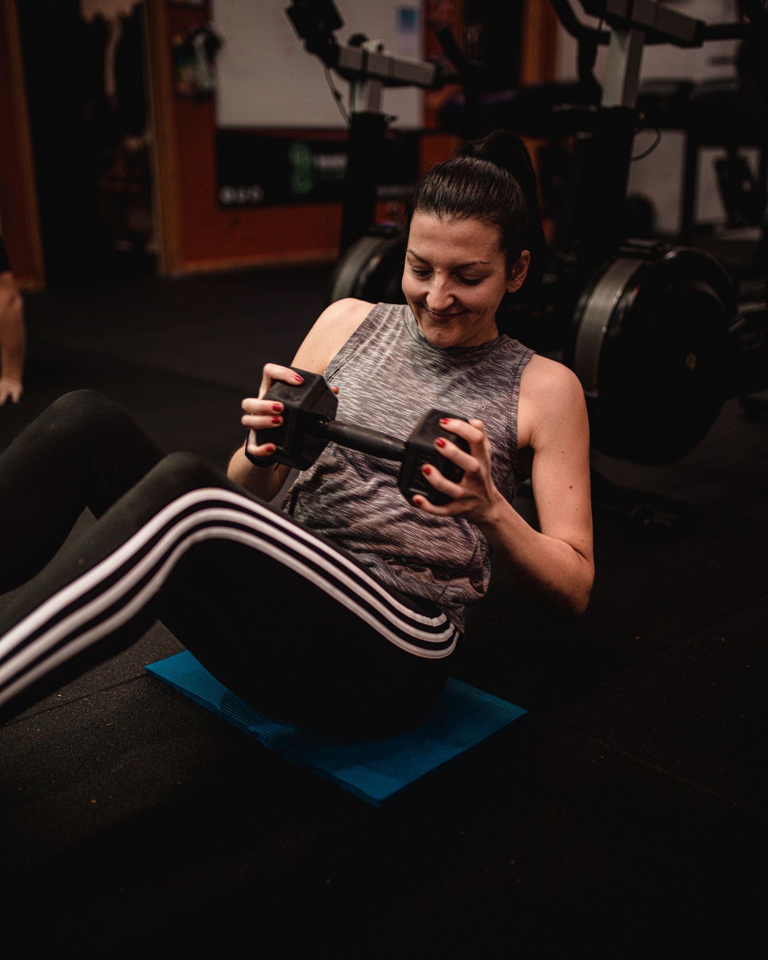 Person in gray tank top and striped pants checking phone while resting on a blue exercise mat in a gym.