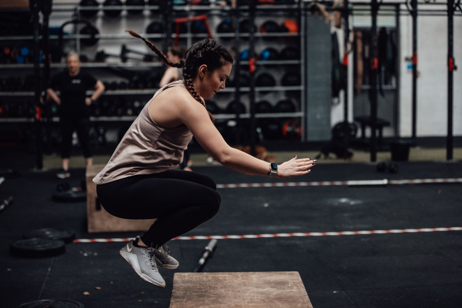 Athlete in beige tank top and black leggings performing squat jump on wooden box in gym.