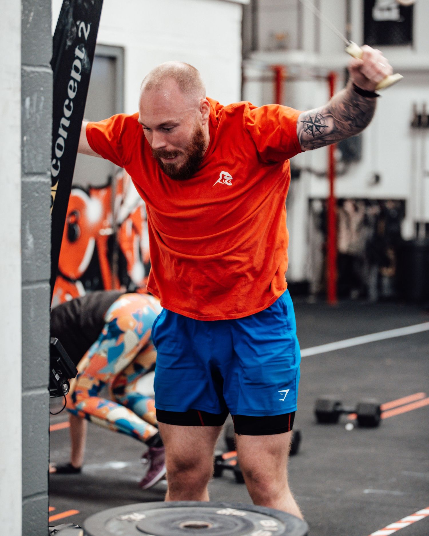 Person in orange shirt and blue shorts performing dynamic exercise movement in CrossFit gym.