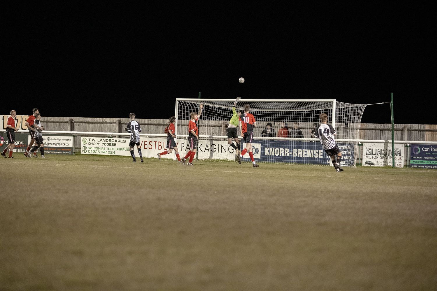 Night soccer match showing players competing near illuminated goal post.