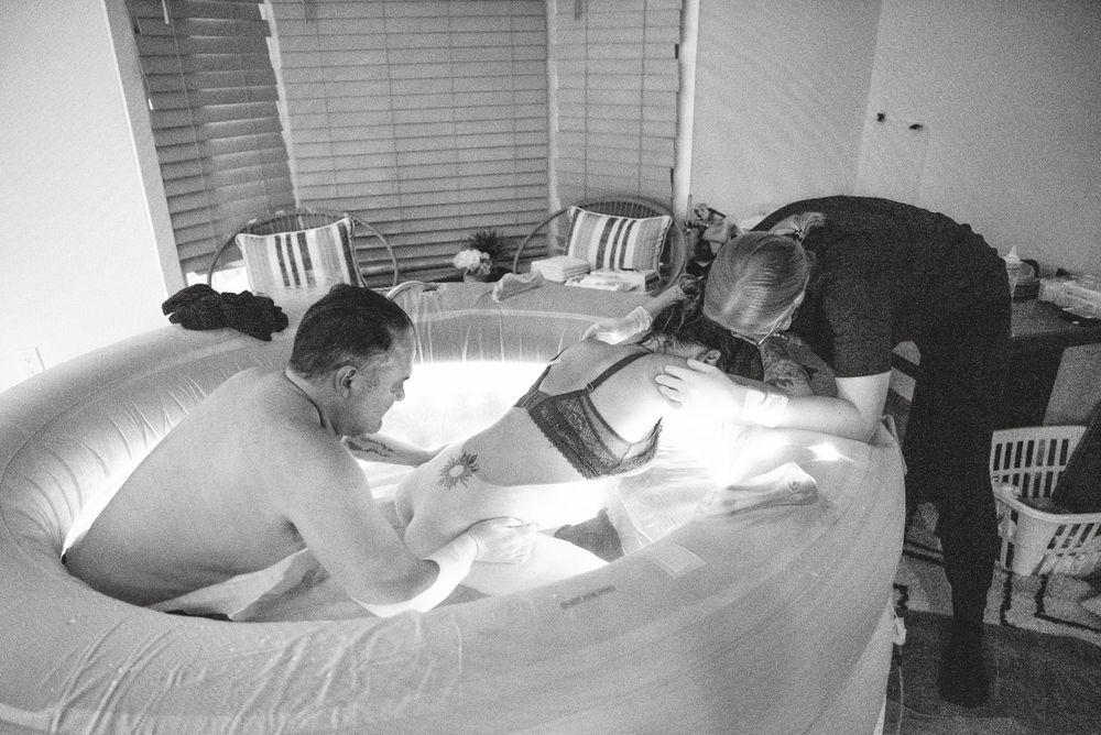 A birthing scene captured in black and white shows support persons attending to someone in a birth pool.