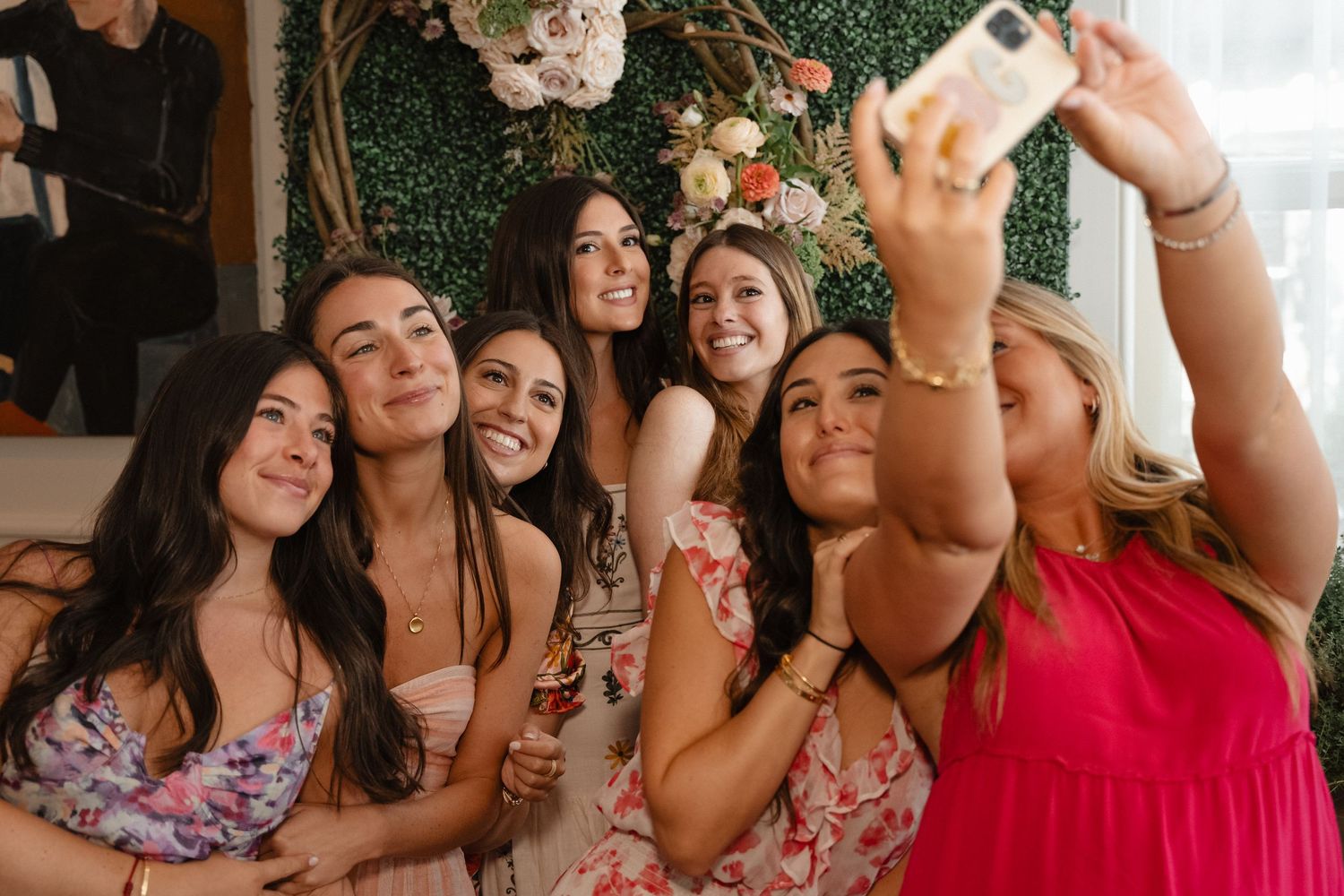 Group of friends taking a selfie together at a wedding celebration with floral wall backdrop.
