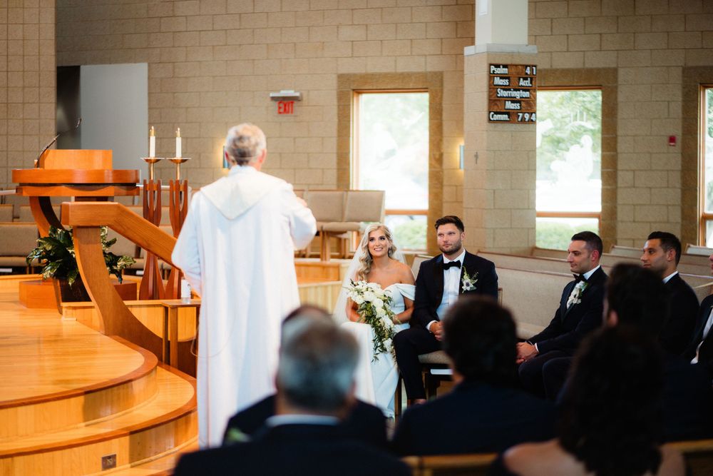 Church wedding ceremony with guests seated in wooden pews and officiating minister in white robe.