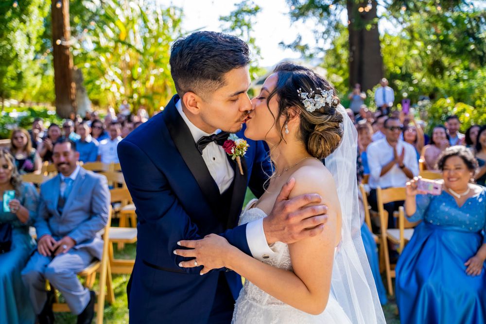 Romantic outdoor wedding ceremony kiss under sunlit tree canopy.