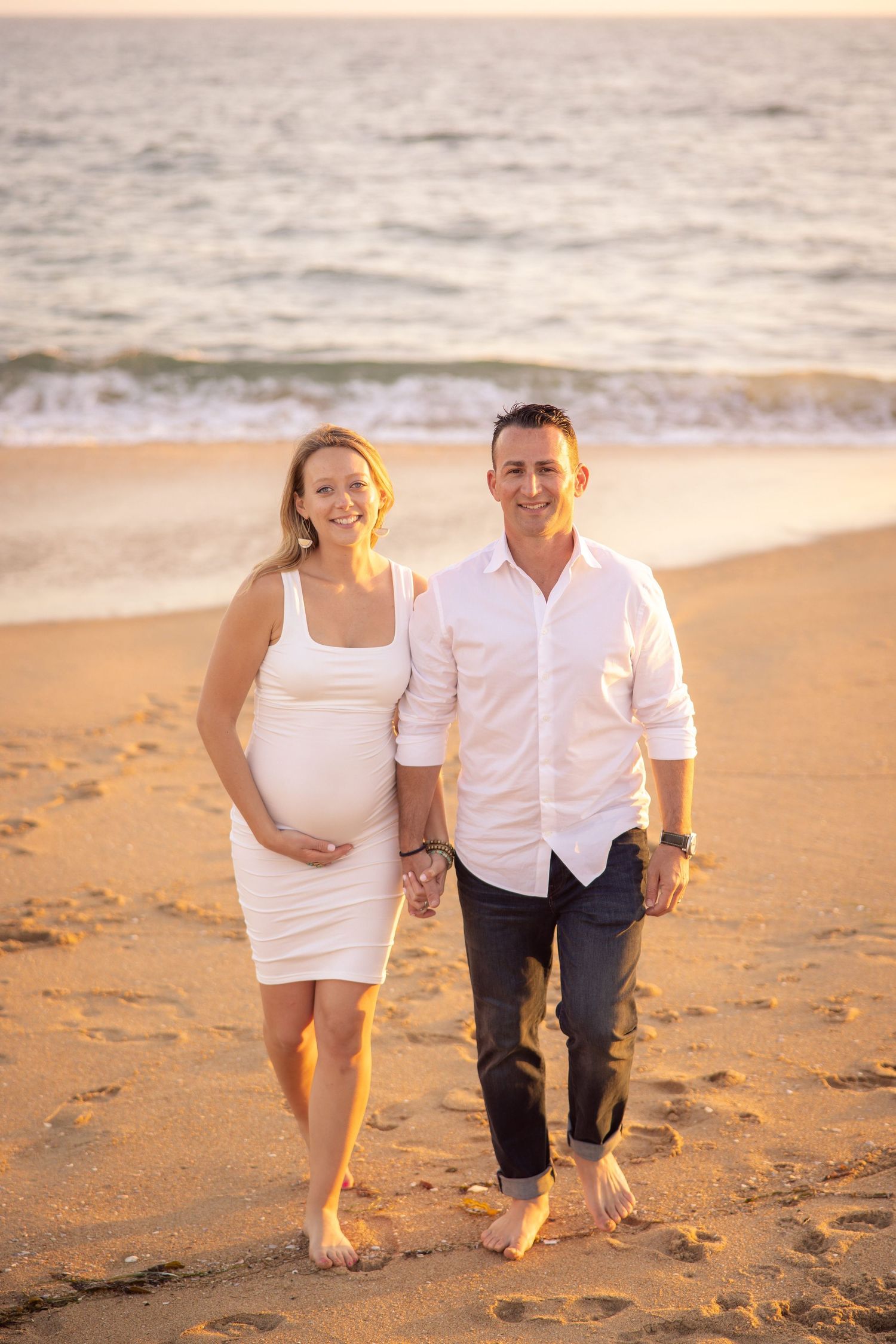 A couple walks barefoot along a sandy beach at sunset during a maternity photoshoot.