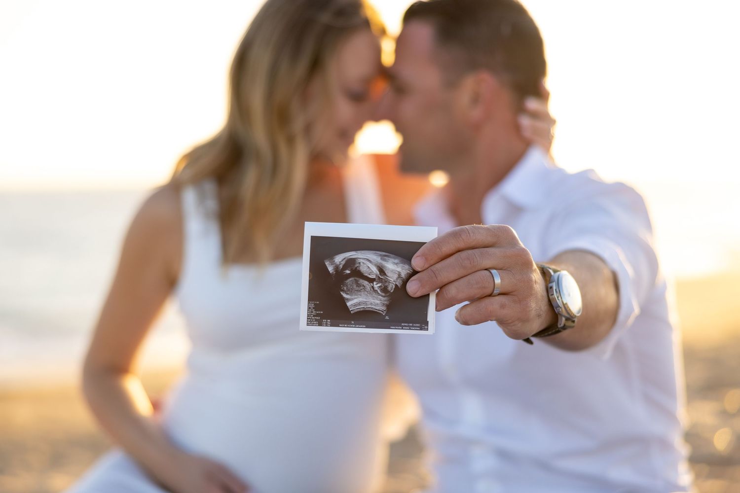 Sunset maternity photo of couple sharing intimate moment while holding ultrasound image.