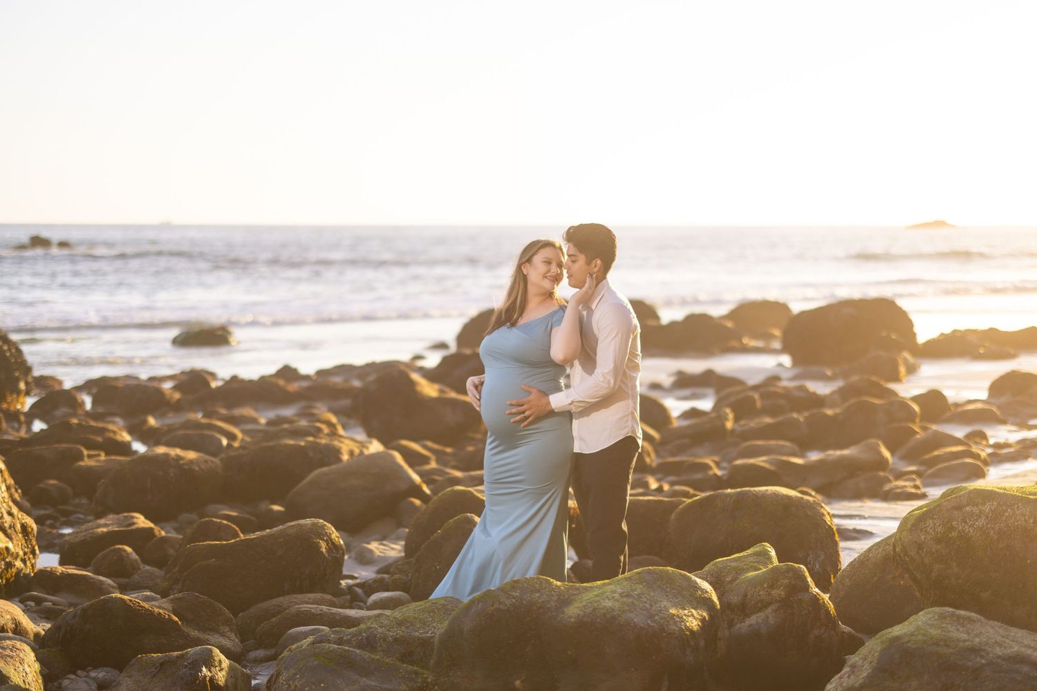 Couple embracing on rocky beach during sunset in formal wear.