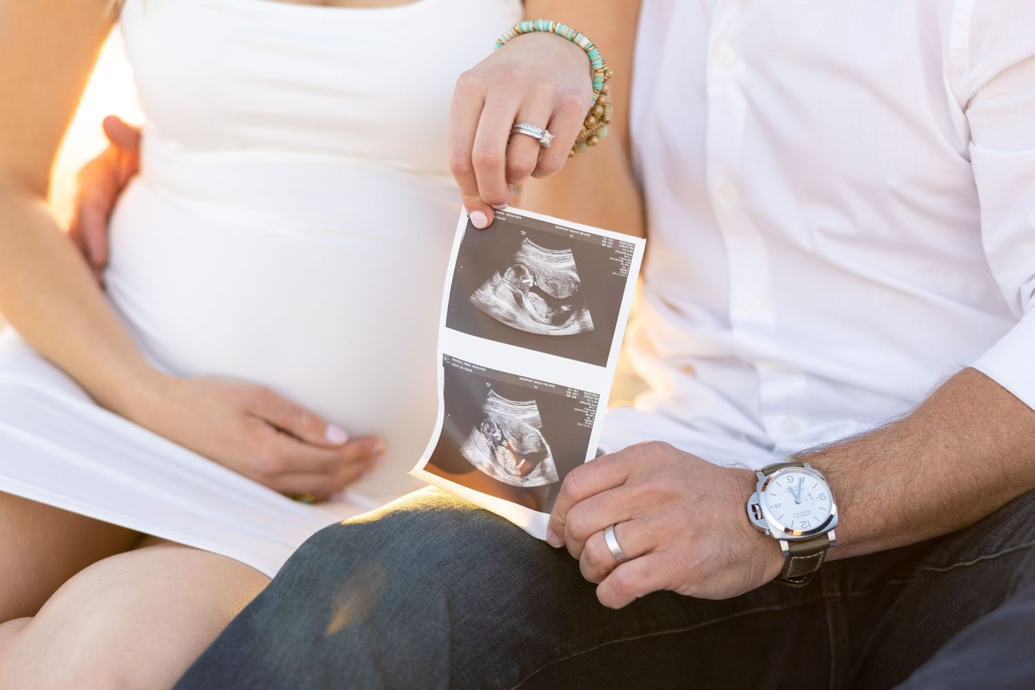 Couple on beach holds ultrasound photos while showcasing pregnant belly in white dress.