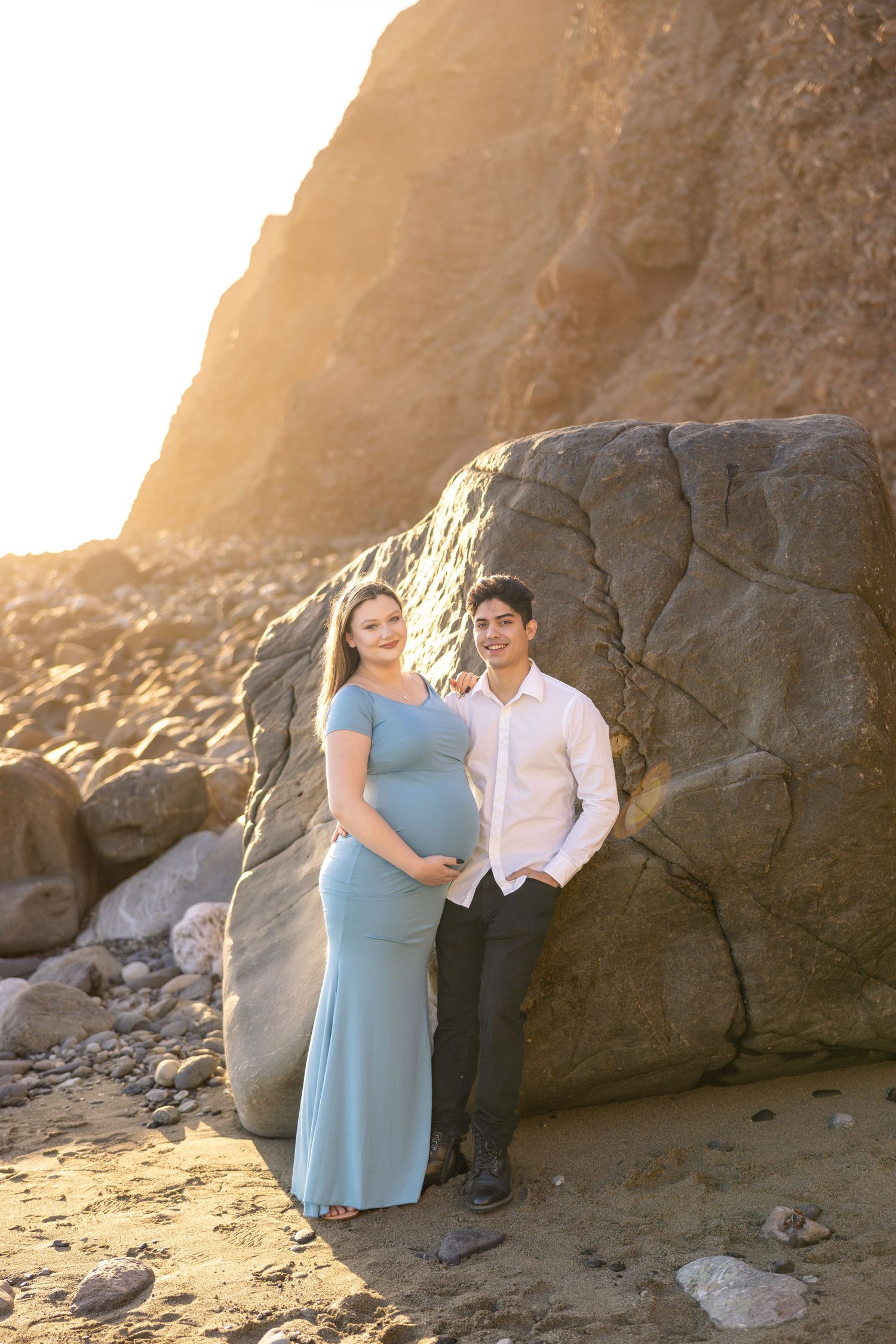 Couple posing by large coastal rocks during golden hour, wearing formal attire.