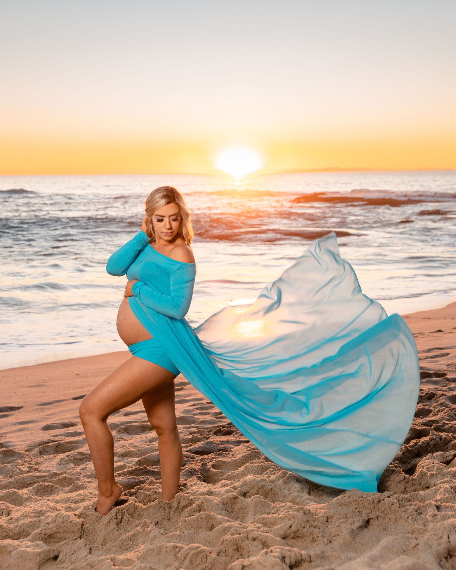 Flowing turquoise dress catching wind during sunset beach maternity photoshoot.
