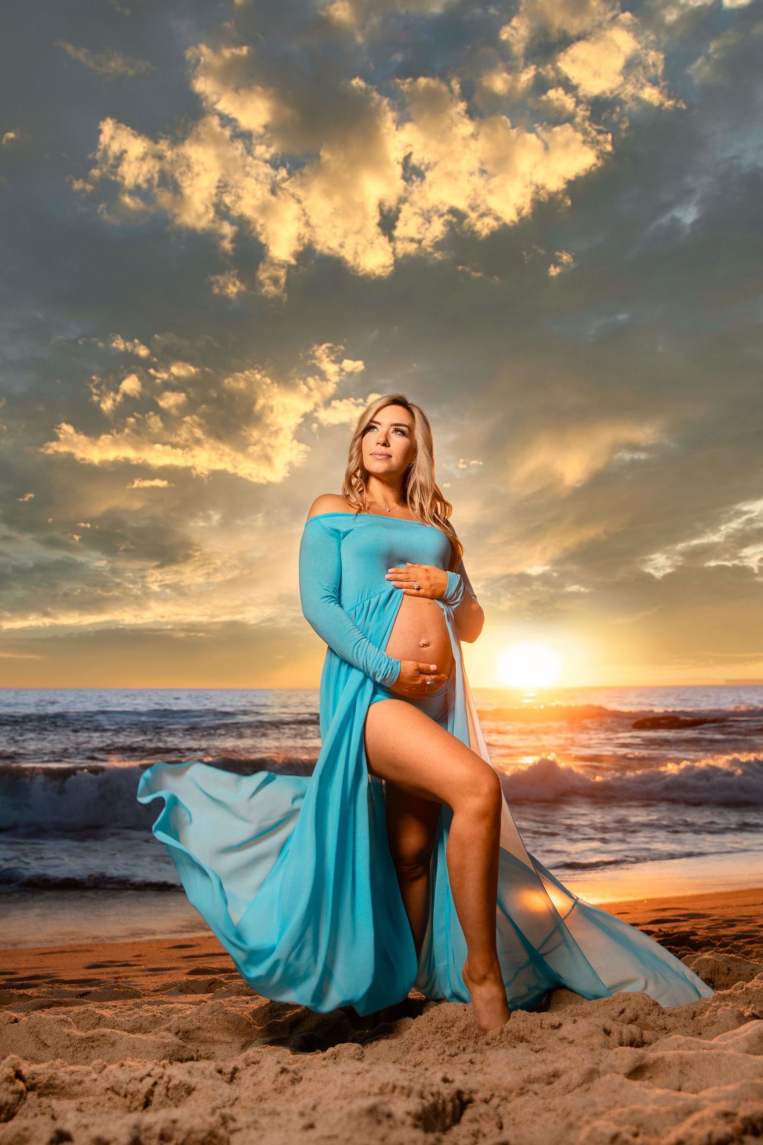 A dramatic maternity photo on beach at sunset with flowing turquoise dress and golden clouds.