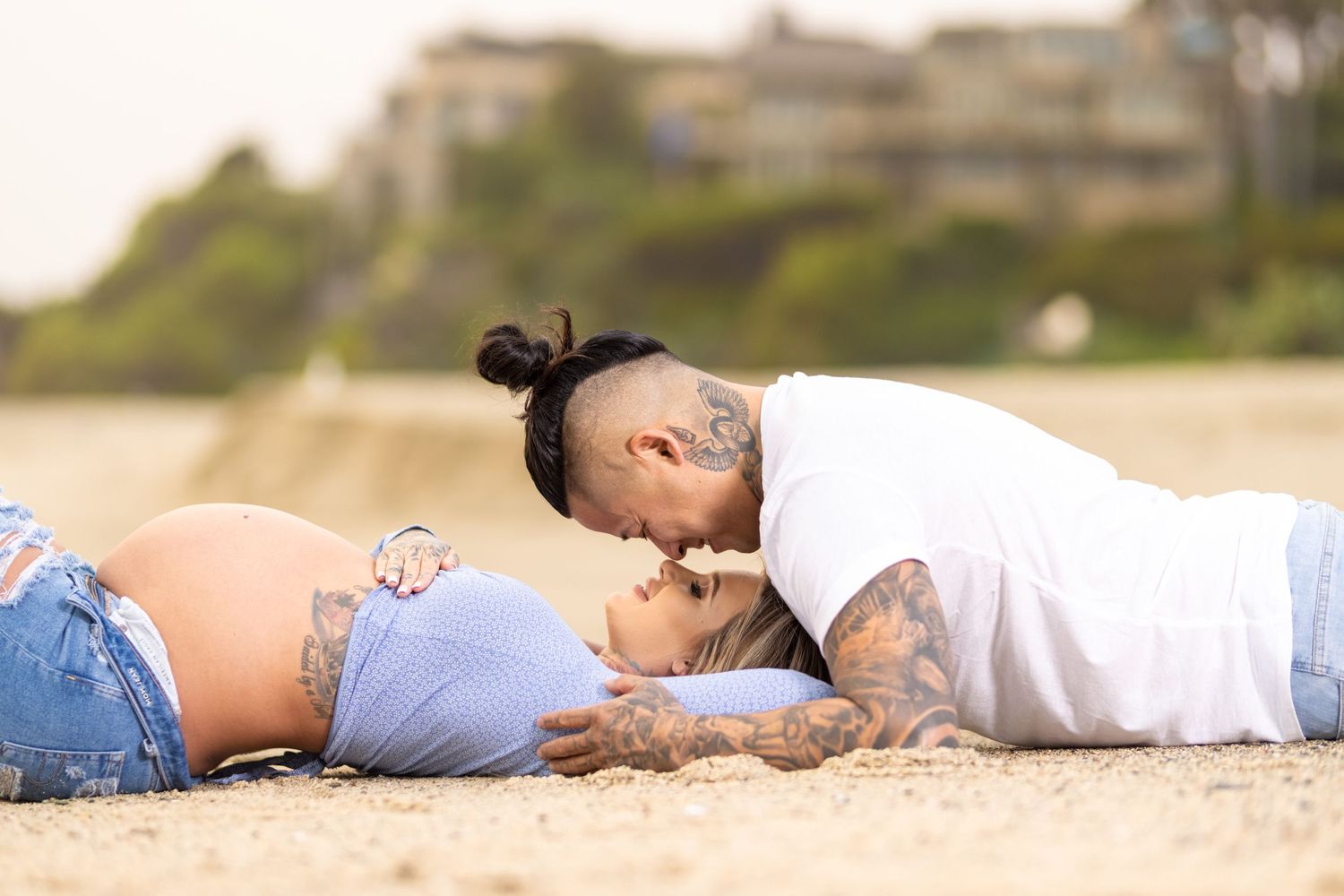 A romantic moment between two people lying on sandy beach with houses visible in background.