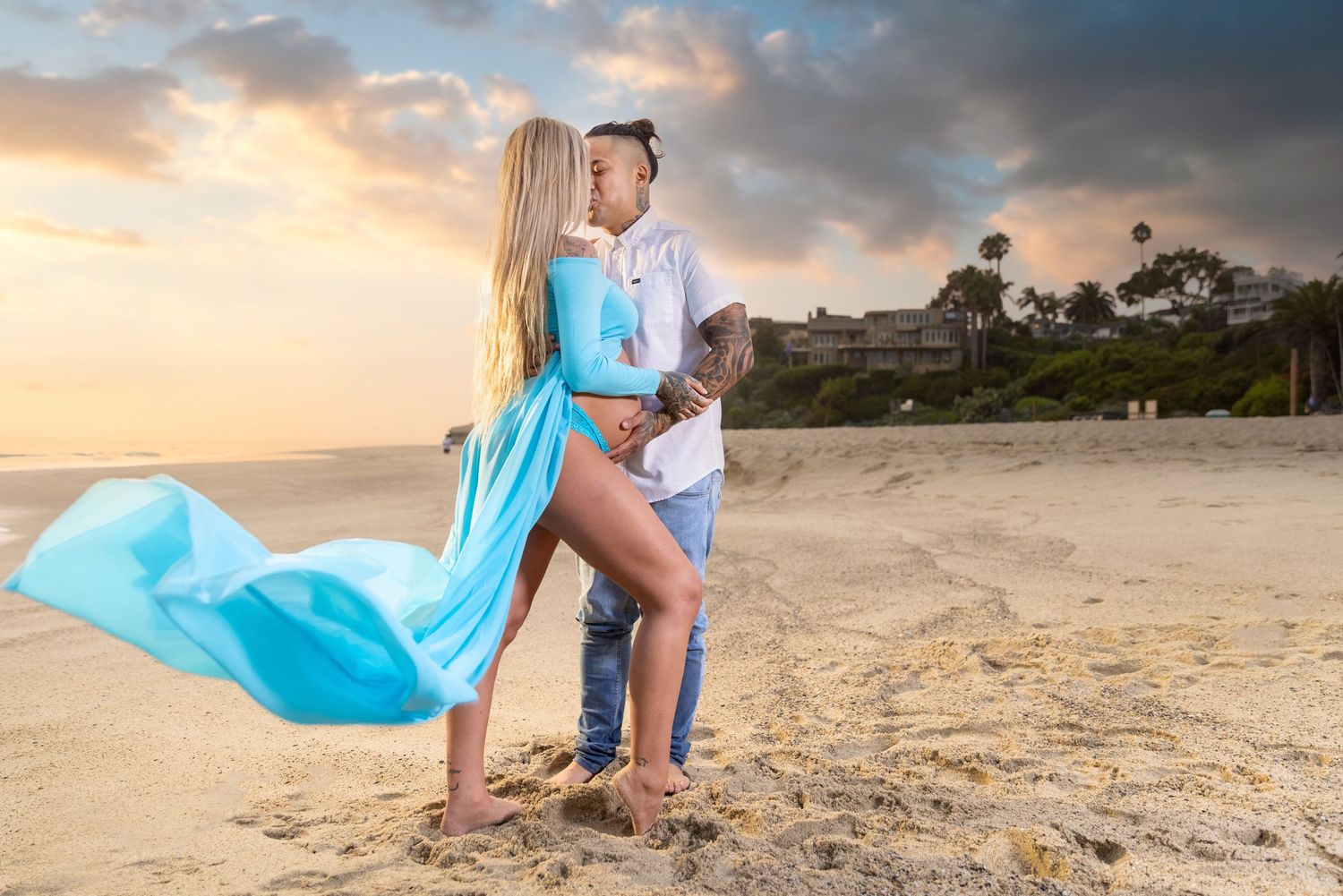 A couple embraces on a beach at sunset with flowing blue fabric catching the wind.