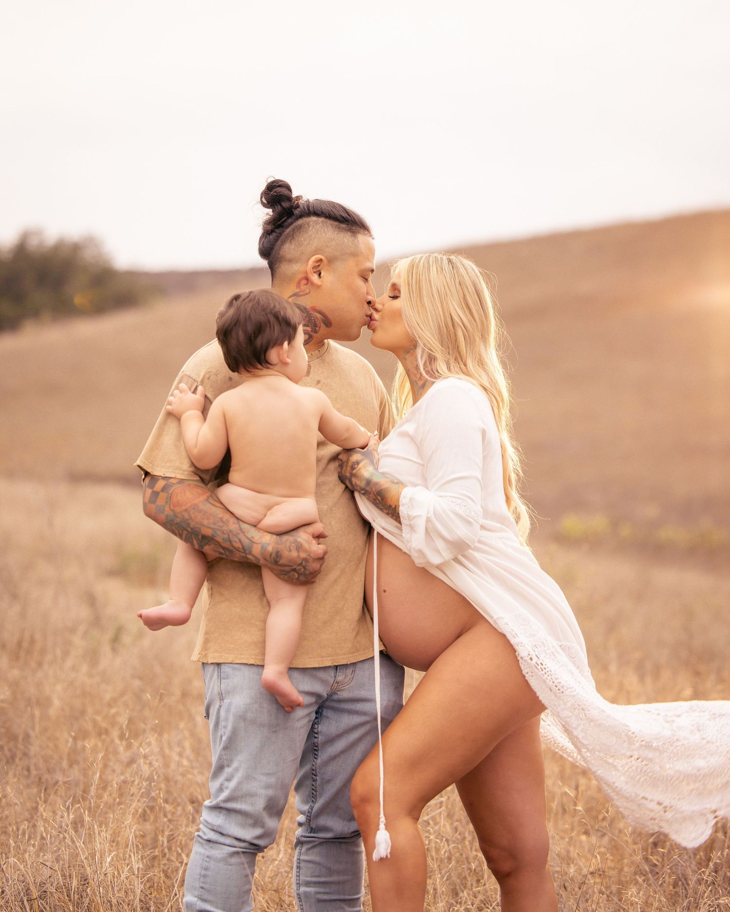 Family sharing tender moment in golden field at sunset with flowing white fabric.