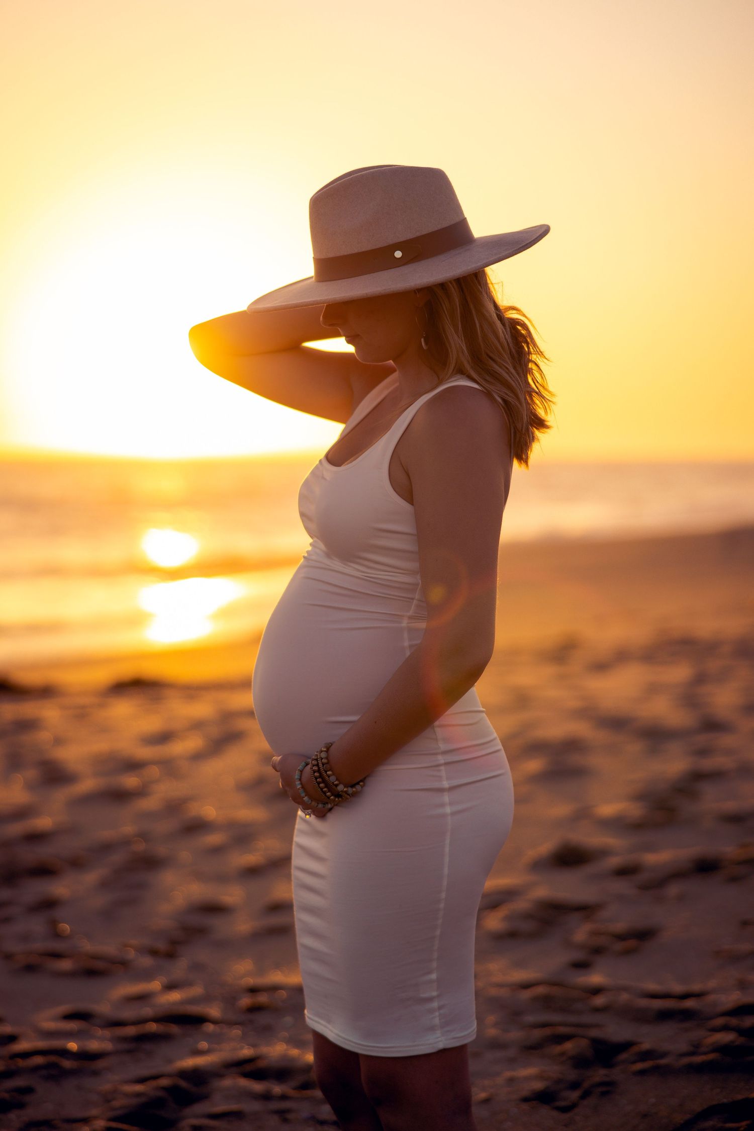 A silhouette of an expectant mother in a white dress and hat poses on a beach at golden hour.
