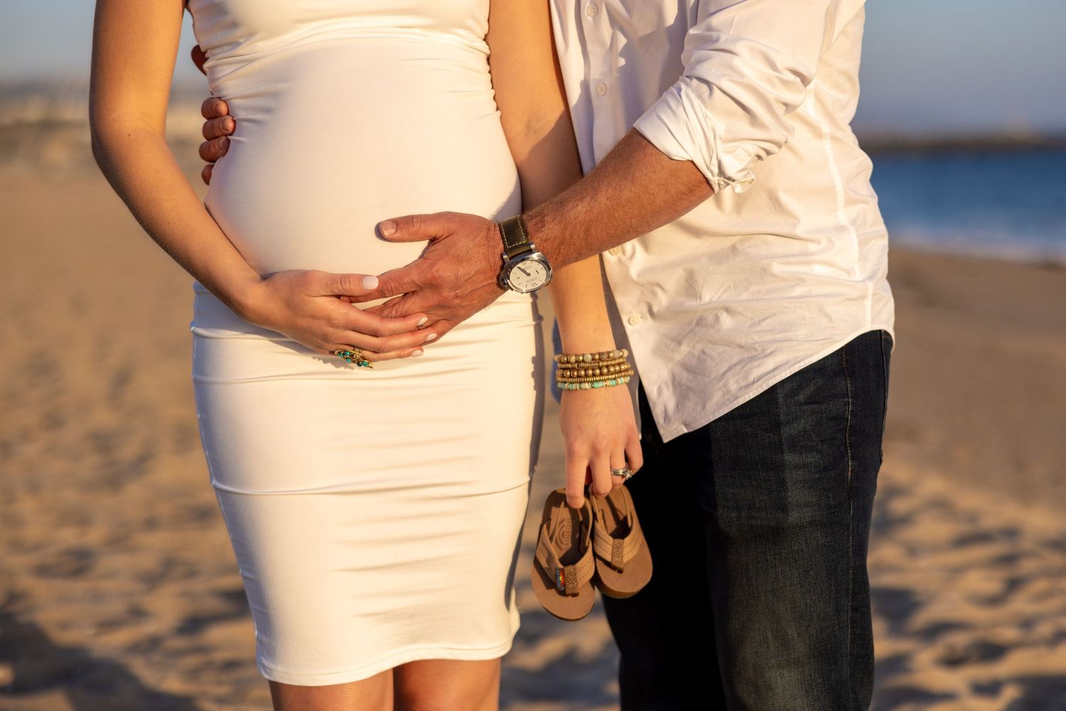 Expectant couple holds hands on beach, focusing on pregnant belly in white dress.