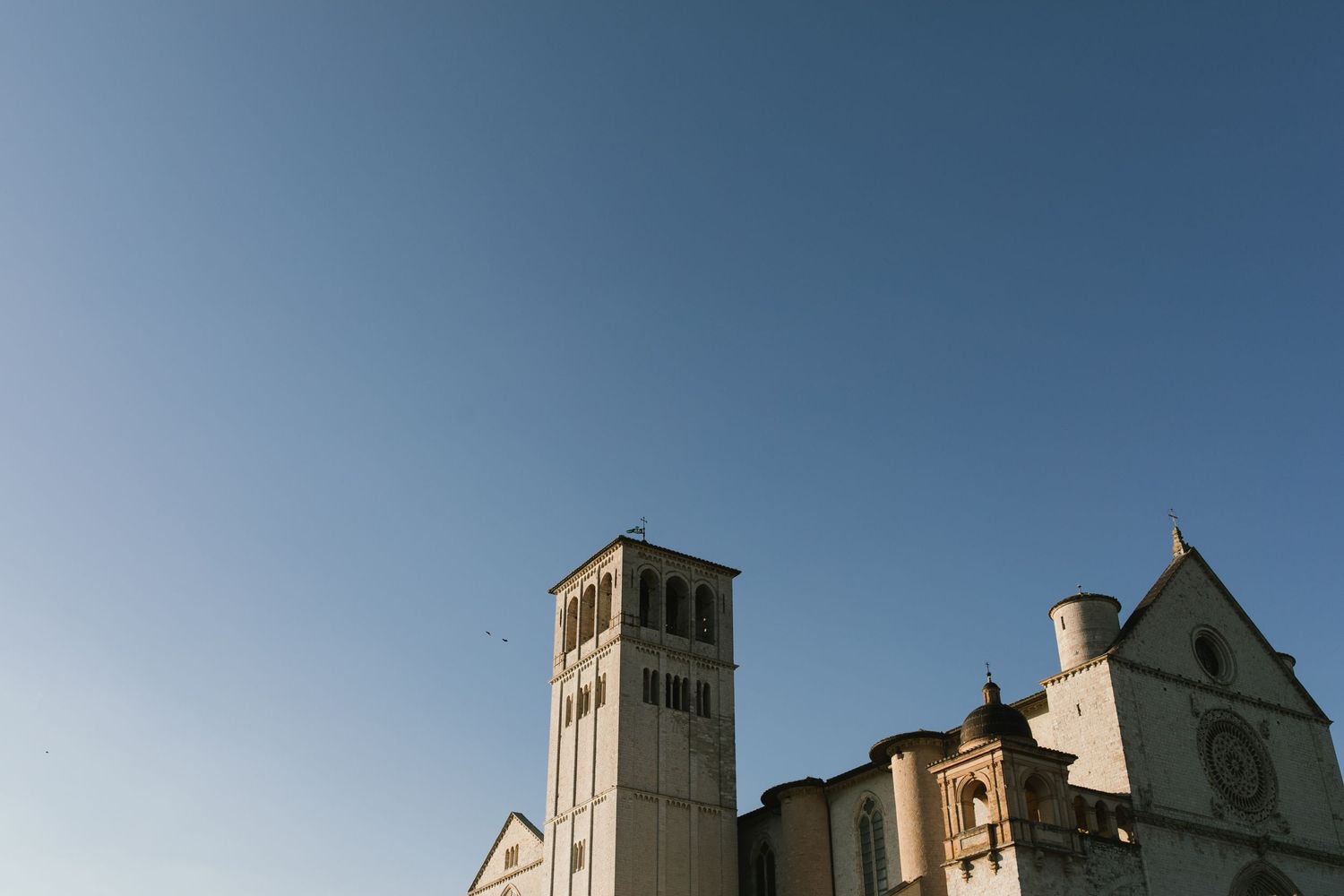 Medieval stone church with tall bell tower against blue sky in Assisi, Italy.