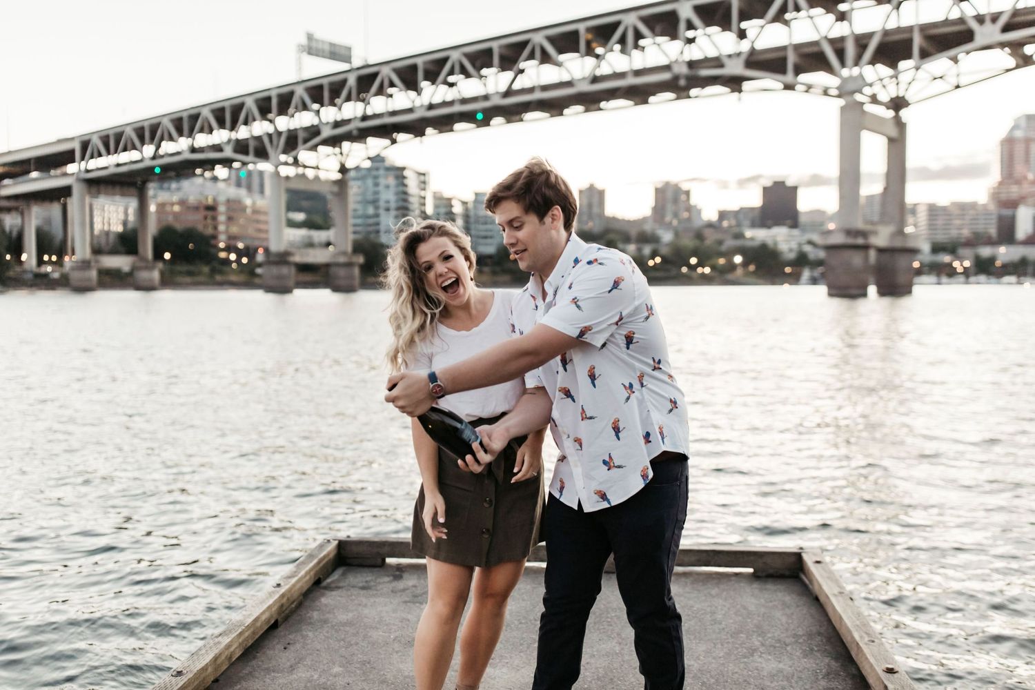 Girl and boy in white popping a bottle of champagne on a dock in front of downtown Portland