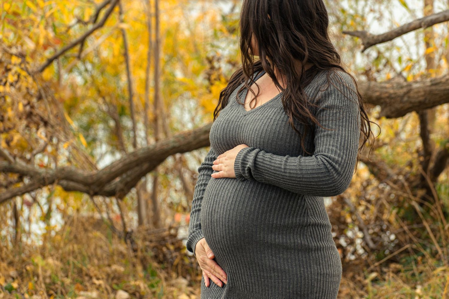A side view of an expecting mother cradling her belly outdoors among autumn colors.