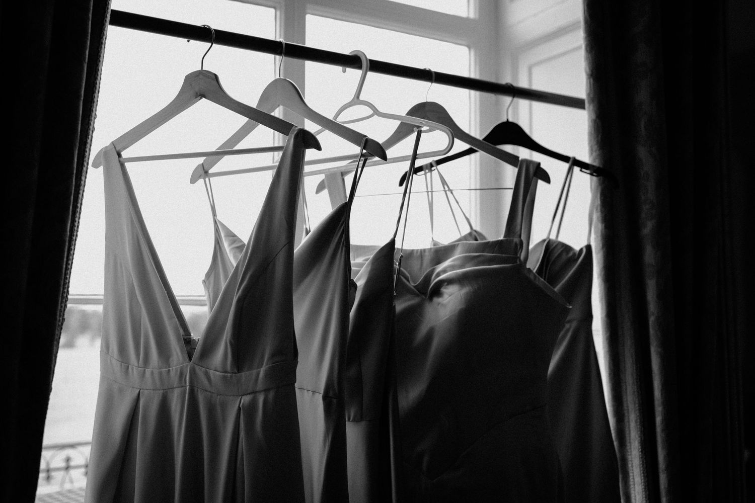 Black and white photo of bridesmaid dresses hanging on wooden hangers by window.