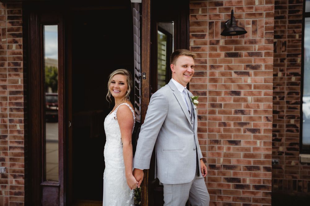 A couple in wedding attire holds hands while standing back-to-back against a brick wall exterior.