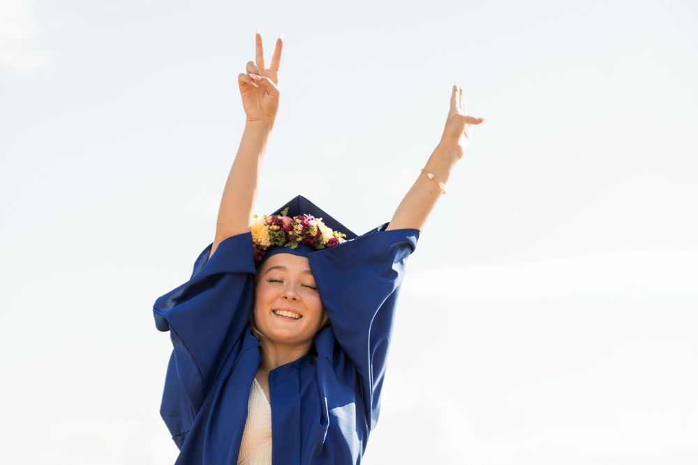 Graduate in blue cap and gown celebrates with victory signs against sky.