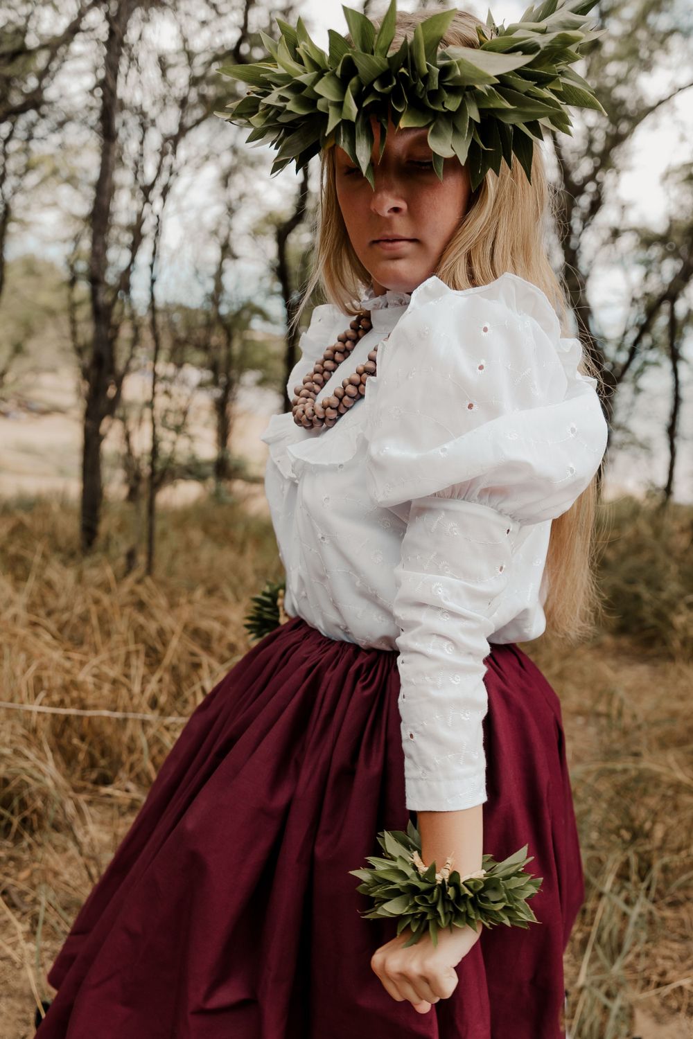 Traditional hula dancer wearing white blouse and burgundy skirt with leaf crown and accessories.