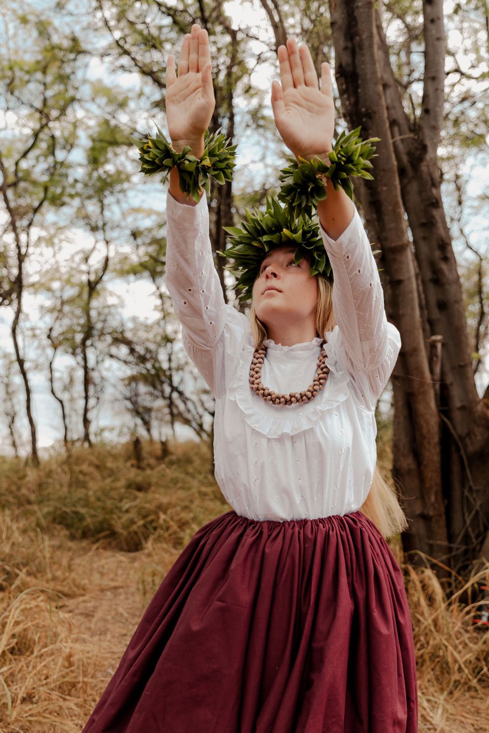 Dancer in traditional Hawaiian attire with lei crown raises arms in forest setting.