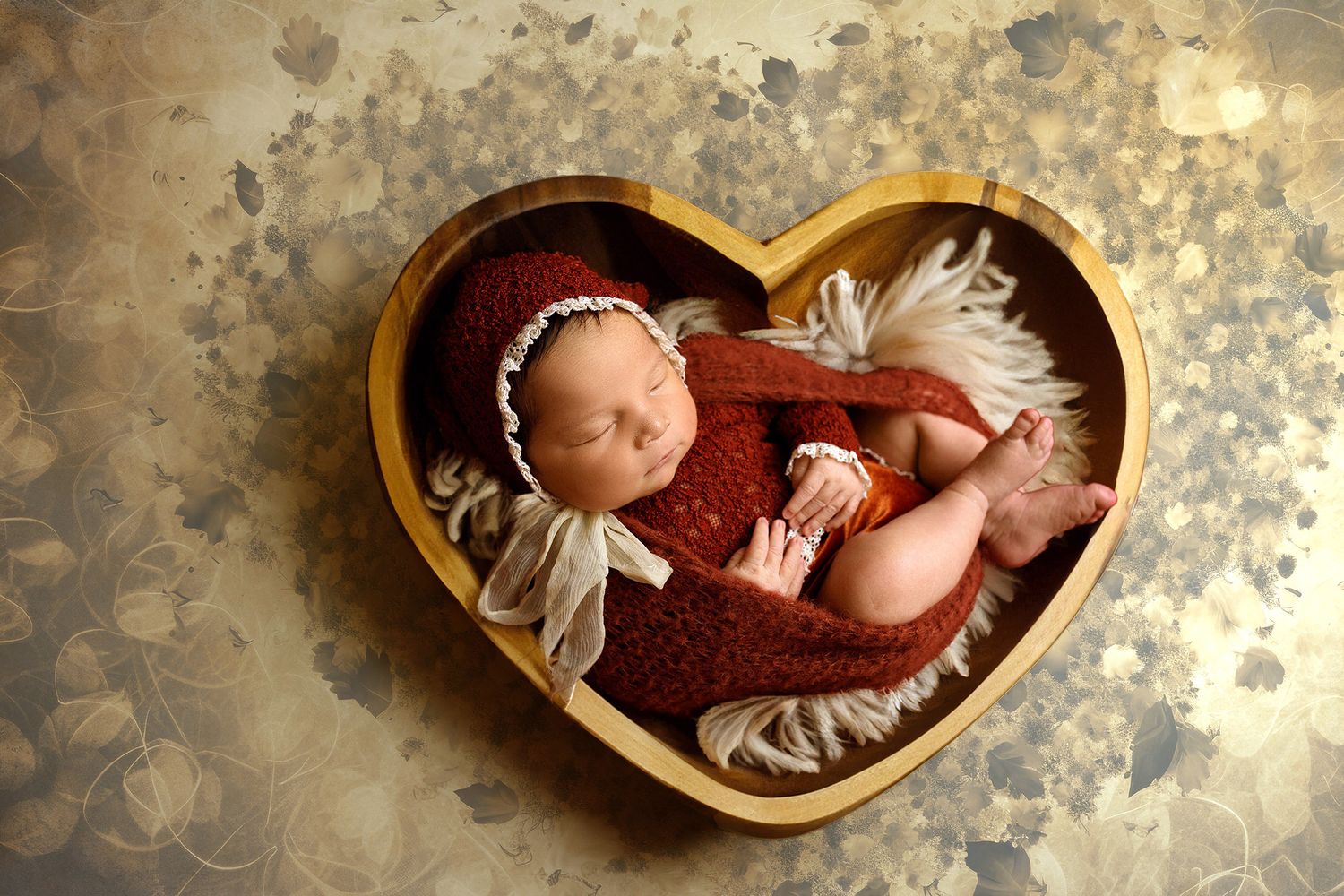 A sleeping newborn in a brown knitted outfit lies peacefully in a heart-shaped wooden bowl lined with soft fabric.