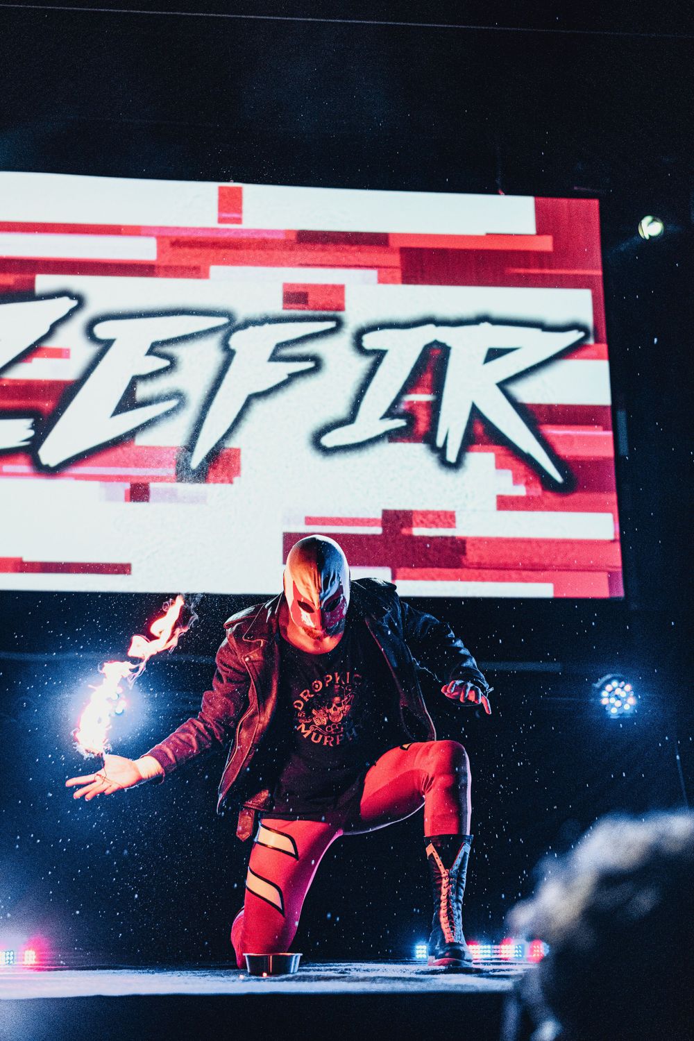 A masked wrestler in red attire performs on stage with dramatic lighting and a red logo backdrop.