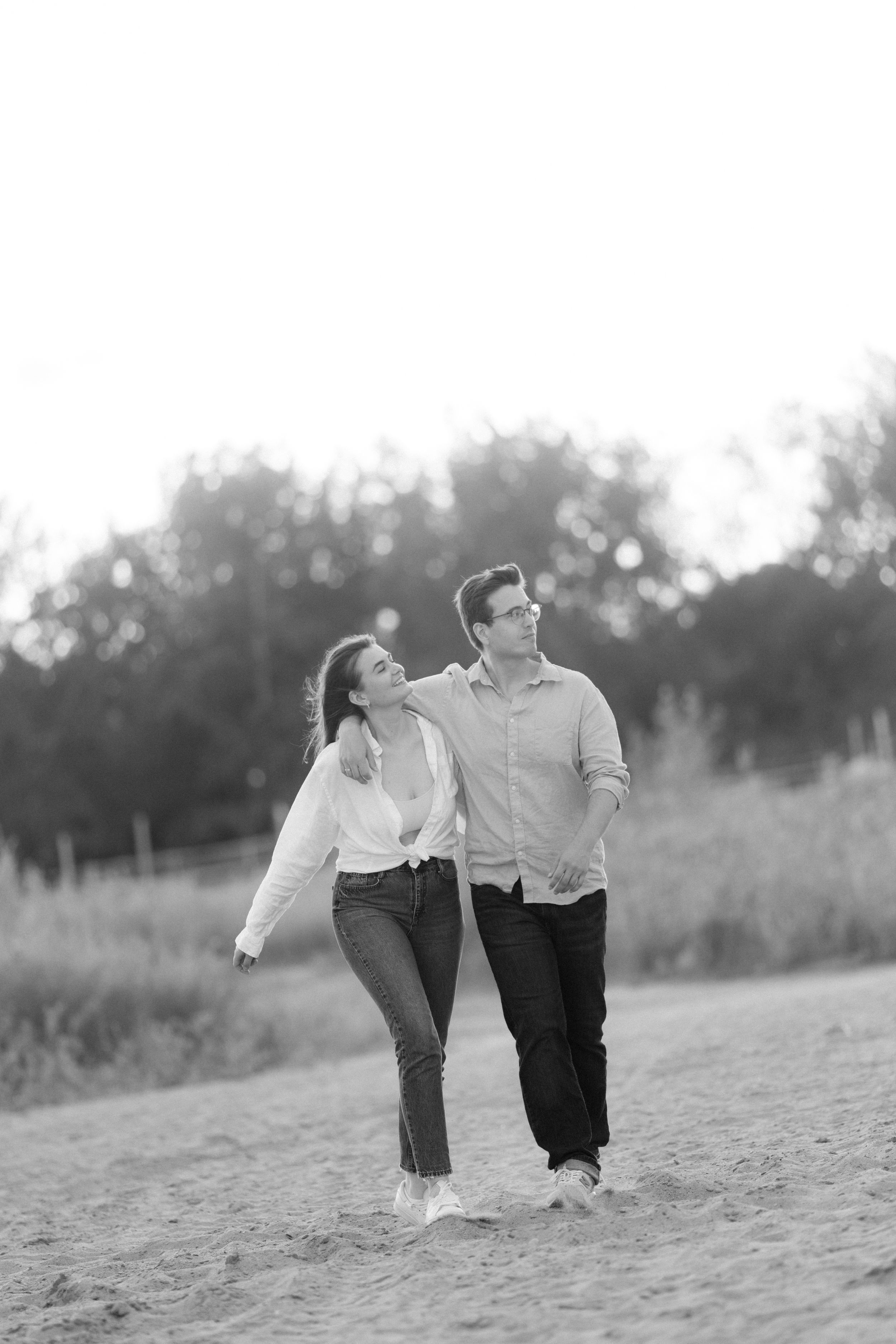 Couple walks together on sandy path during black and white sunset photoshoot.
