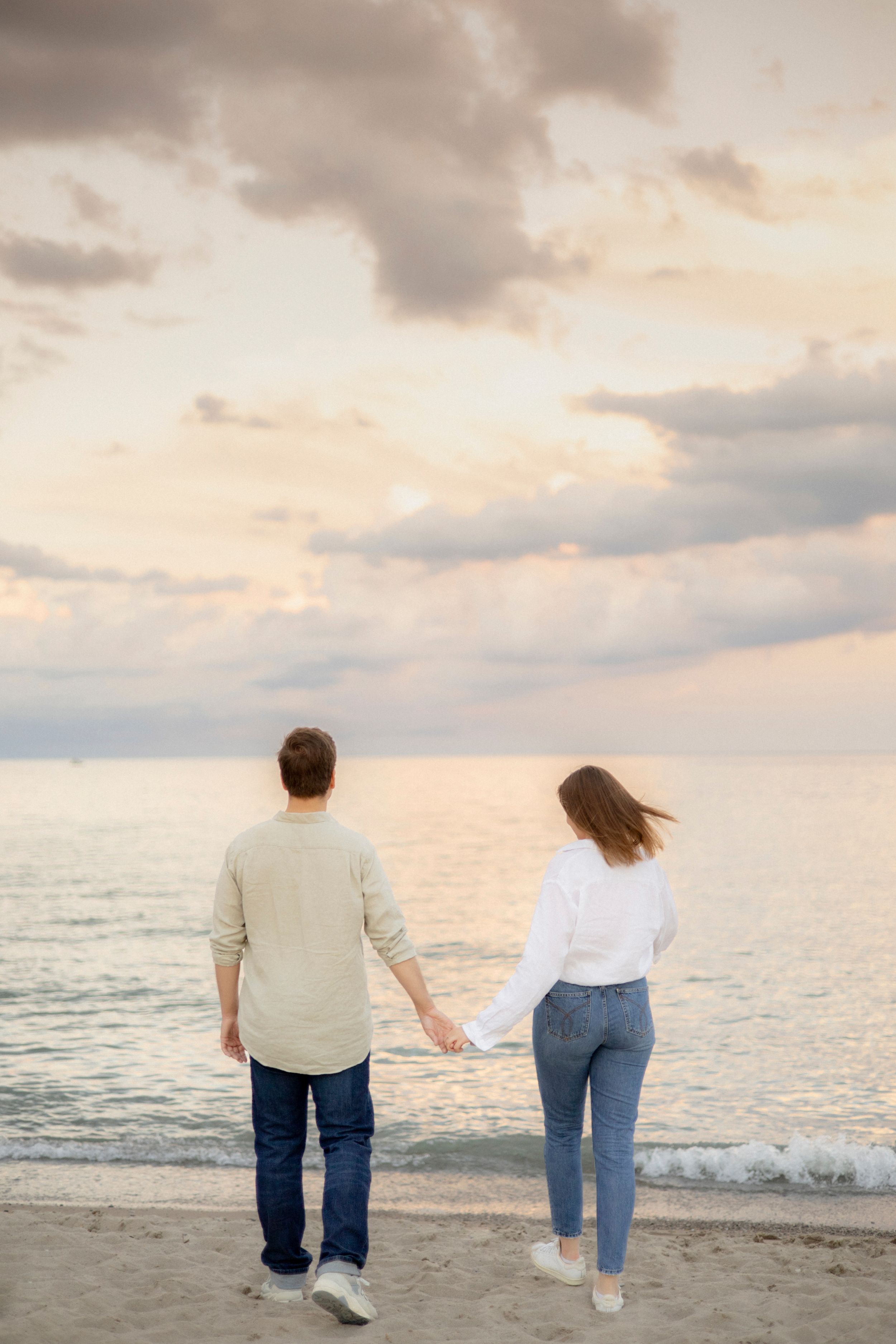 A couple holds hands while walking on the beach at sunset with dramatic cloudy skies above.