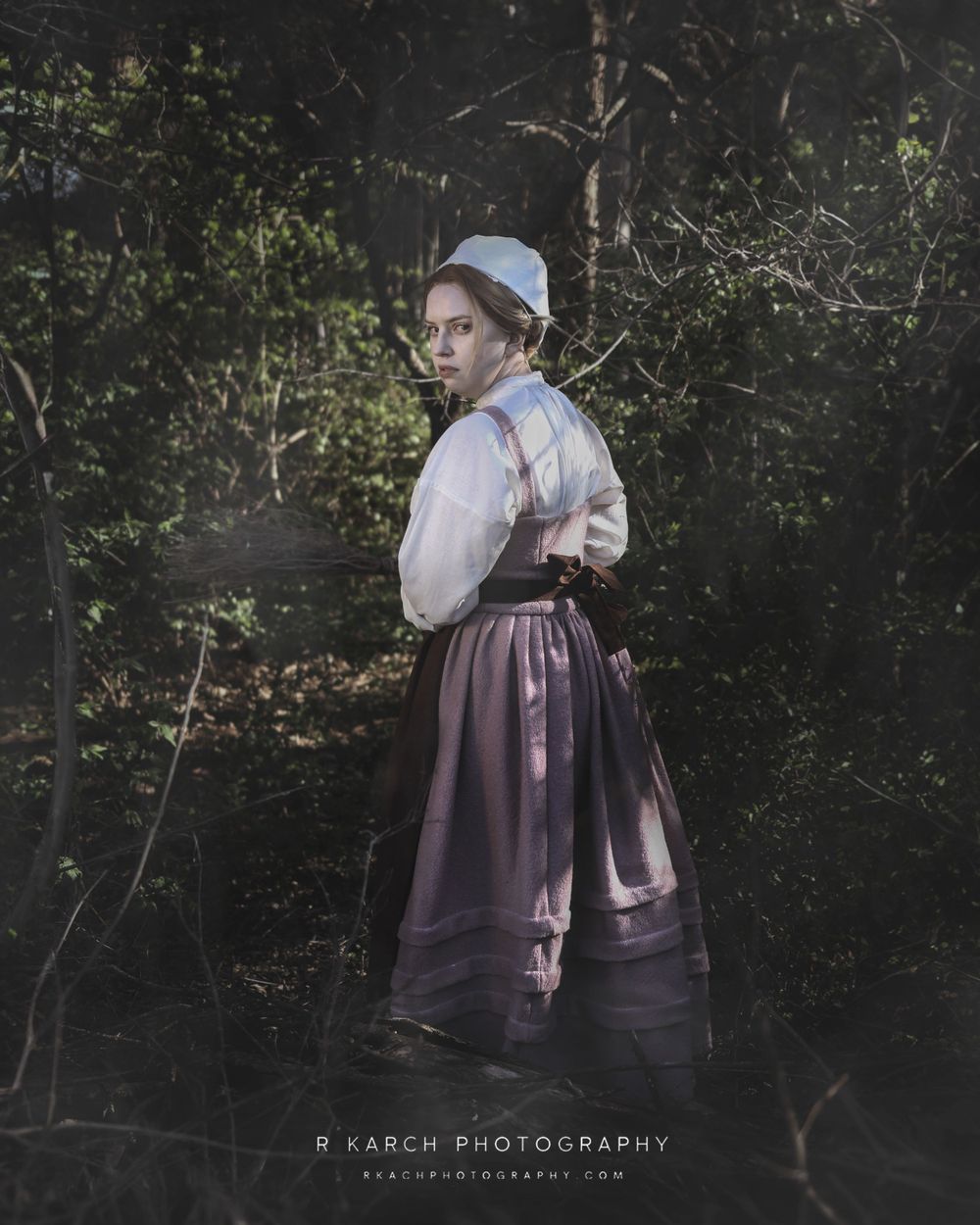 A figure in historical dress poses in a dark woodland setting wearing a bonnet and full skirt.
