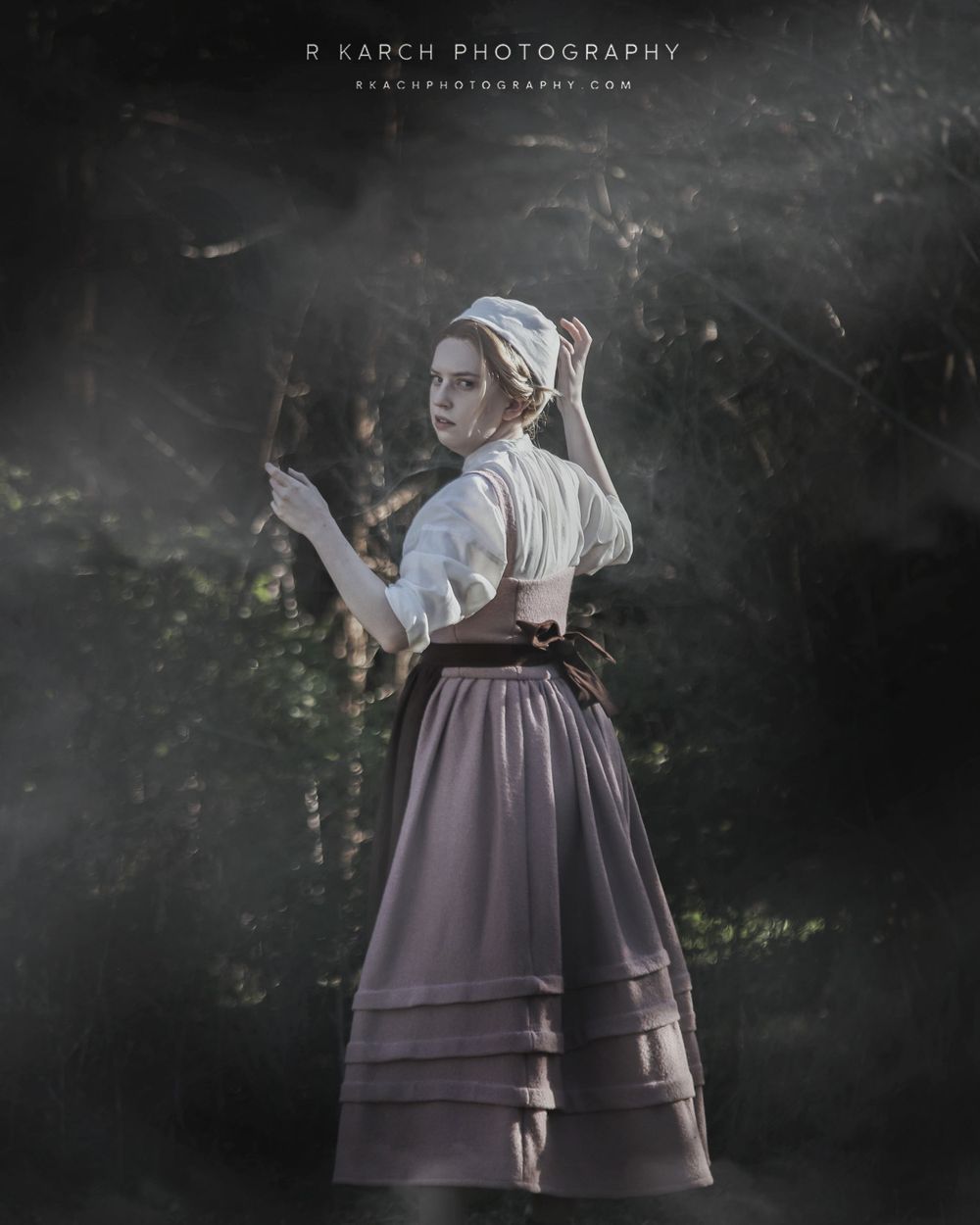 Period costumed figure in a white blouse and dark layered skirt stands in atmospheric moonlit woods.