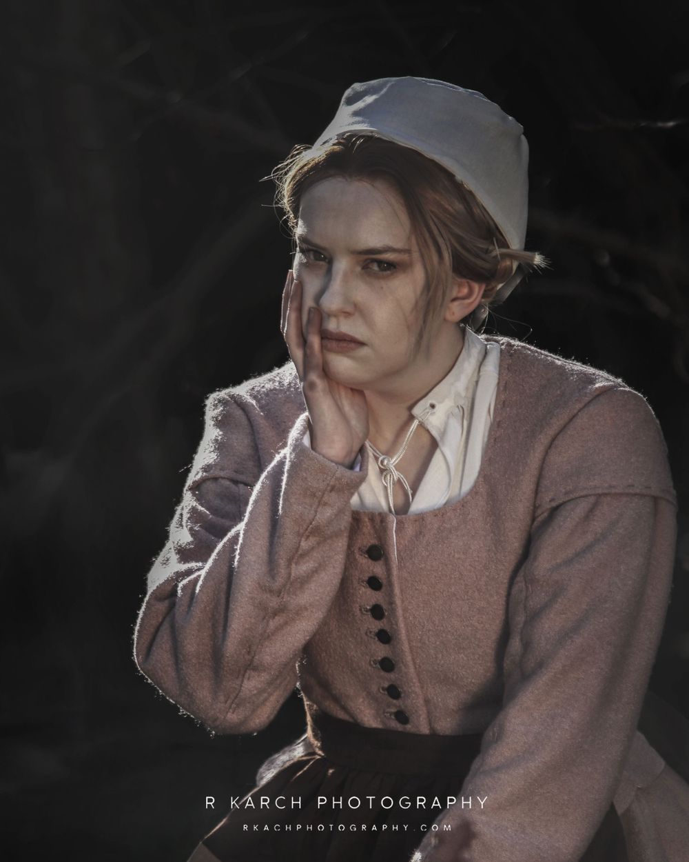 Figure in historical dress with white collar and dusty rose jacket poses against dark background.