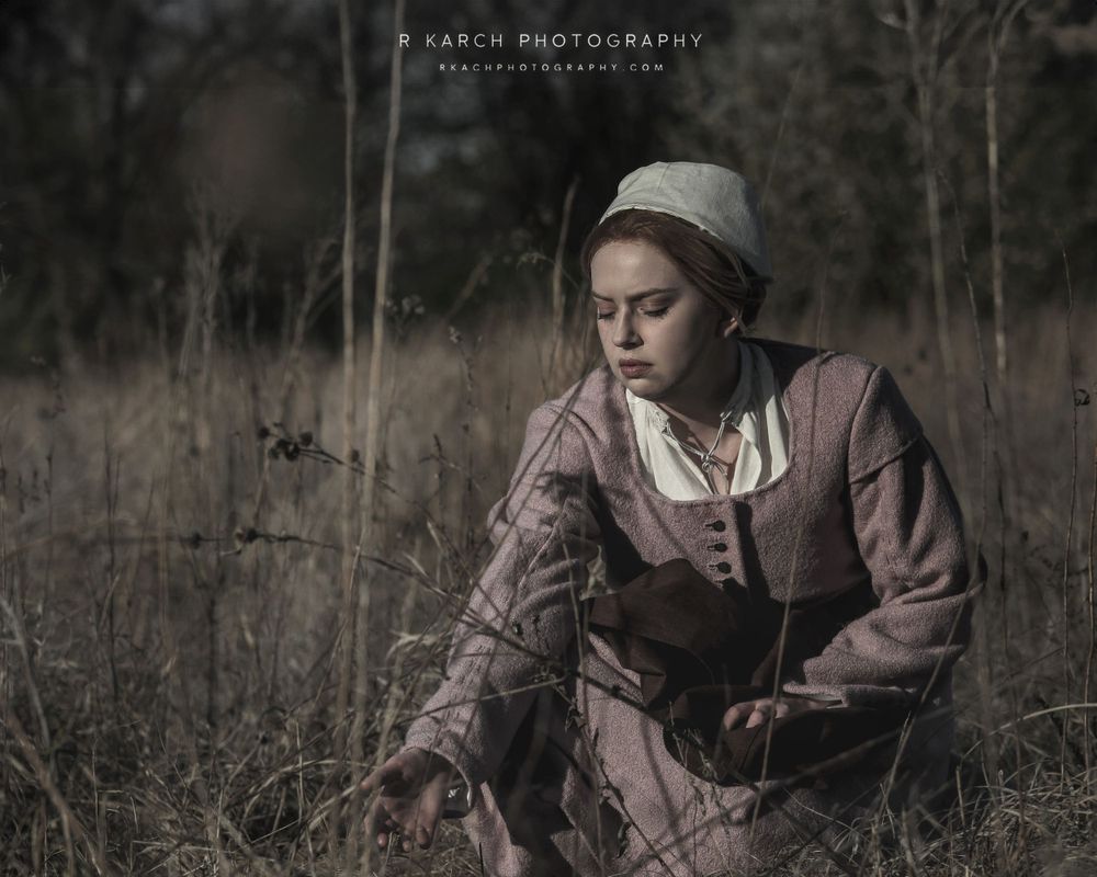 Atmospheric portrait of person in colonial-era clothing sitting in a misty field.