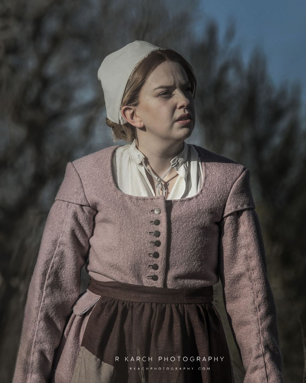 Figure wearing traditional colonial dress with white cap and rose jacket in natural outdoor setting.