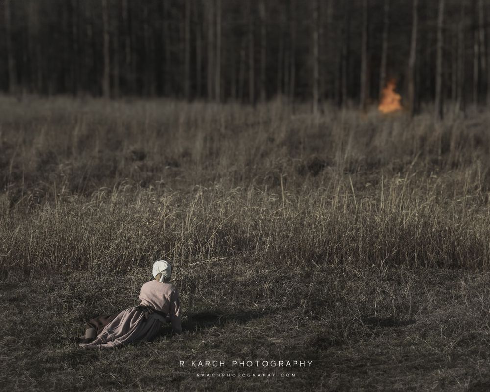 Lone figure in period dress sits in field with distant fire visible through trees.