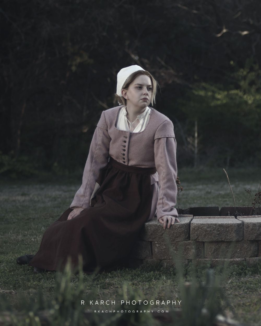 Person in historical clothing sits thoughtfully on stone wall at dusk.