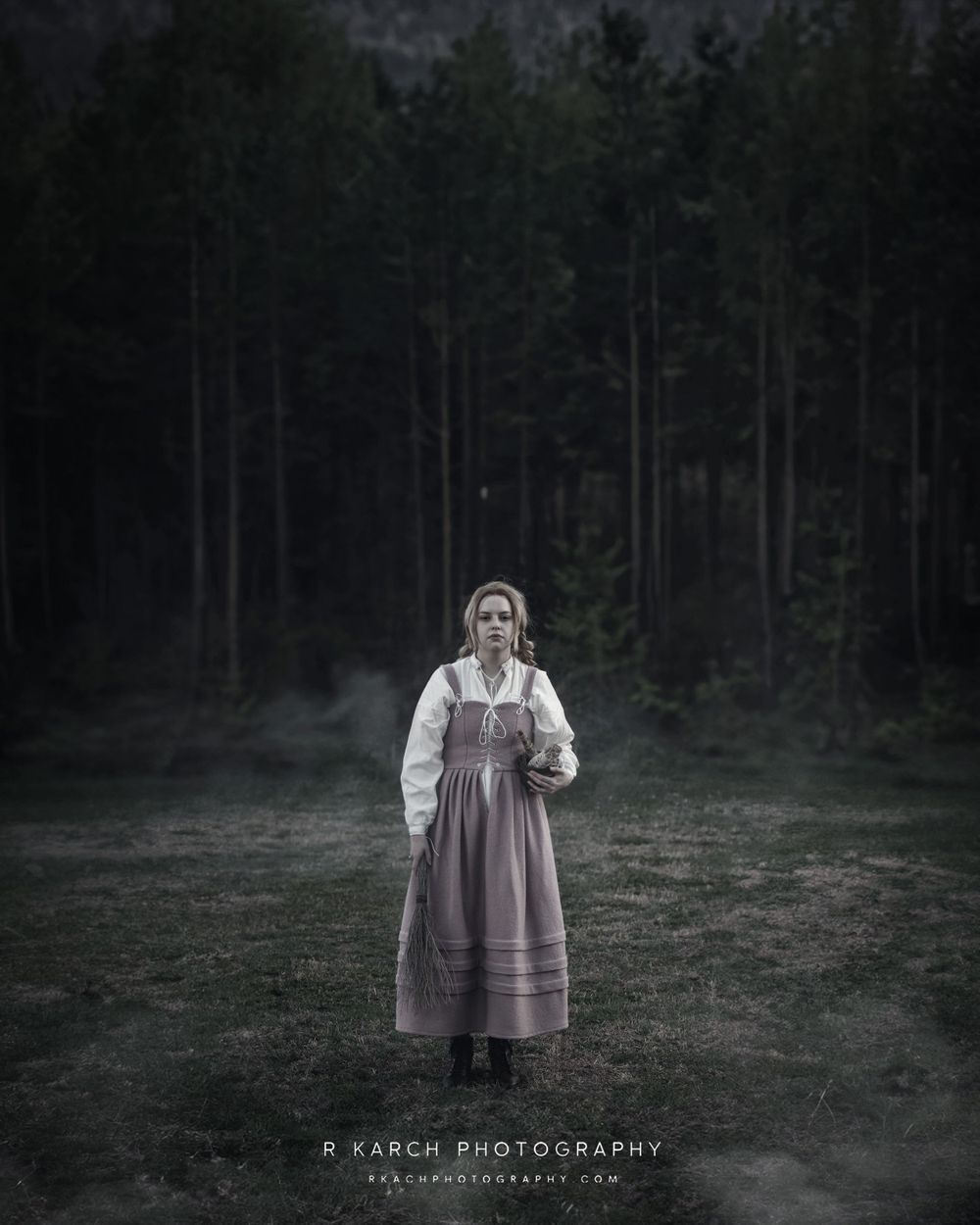 A moody atmospheric portrait in period dress standing in a misty field at dusk with dark forest in background.